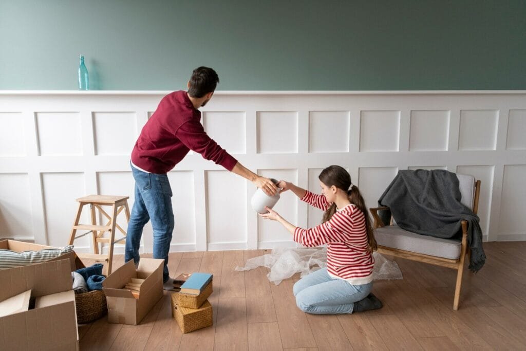 Two people decorating a room, with one standing and handing a lamp to the other who is kneeling by an open box, preparing for move-out cleaning.