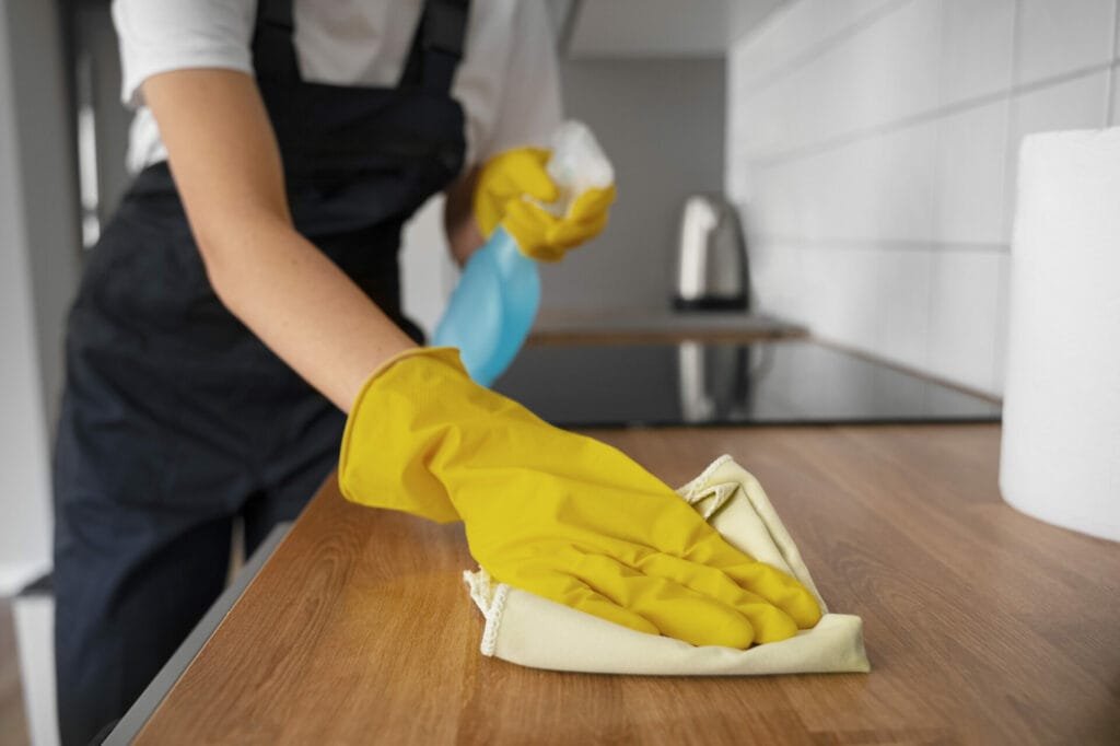 Person in an apron and yellow gloves performing vacation rental cleaning on a kitchen counter with a spray bottle and cloth.